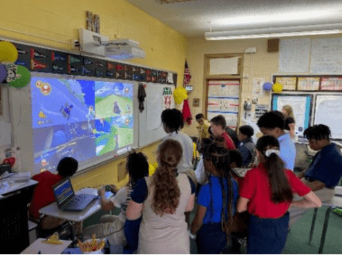 Children and adults watch a video game projected on a whiteboard.
