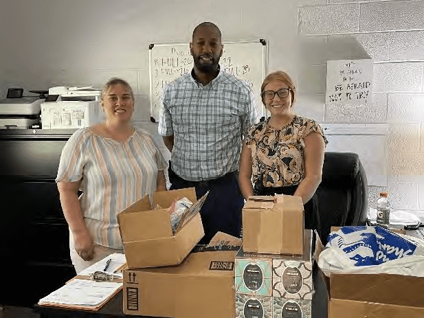 Three adults stand smiling behind a table with boxes.