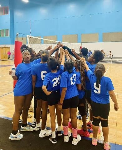 Children in blue shirts huddle on a volleyball court.