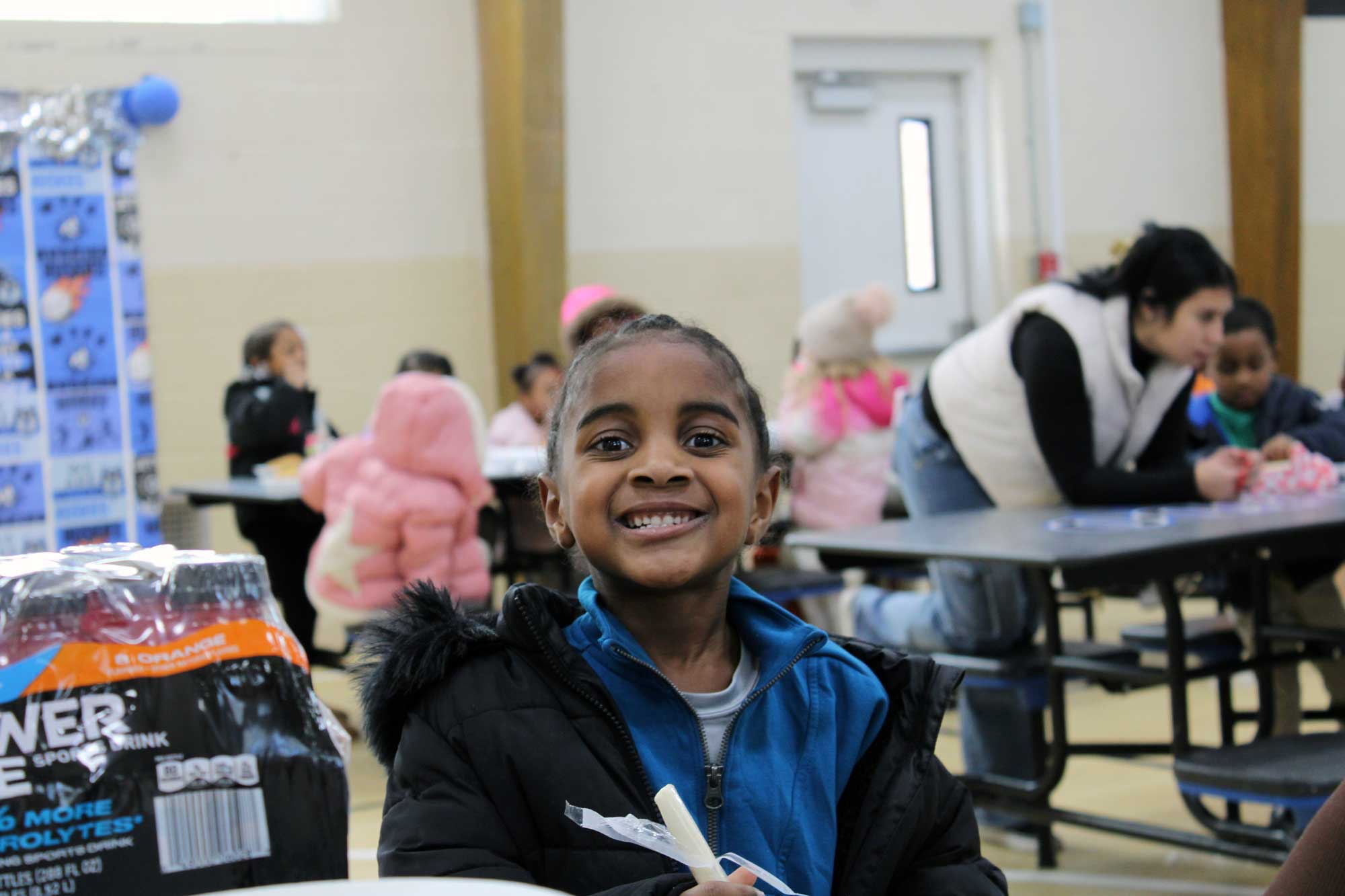 Smiling young student holding a snack at a table with classmates and staff behind.