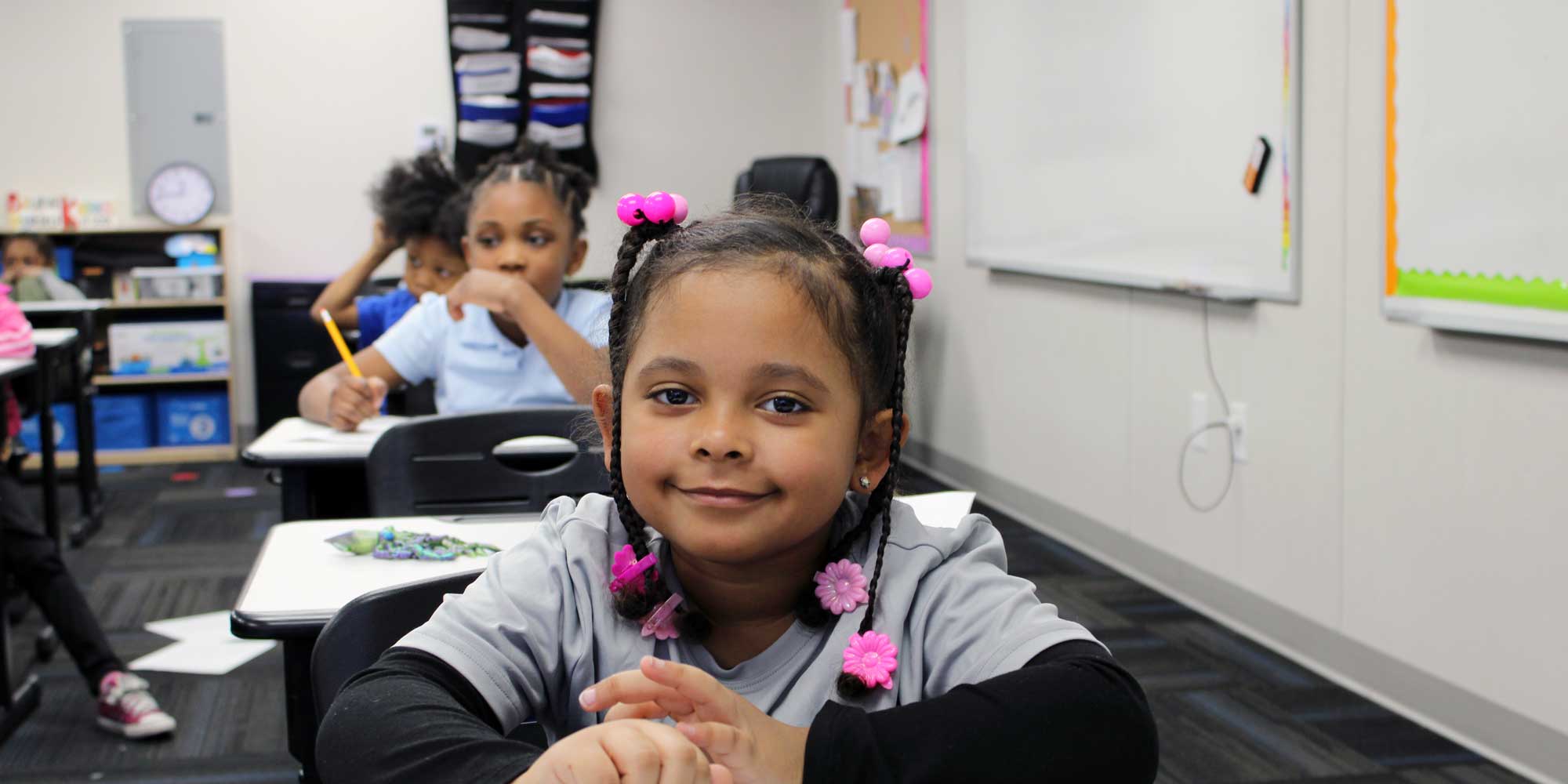 Smiling young student sitting at a desk in a classroom with classmates behind.