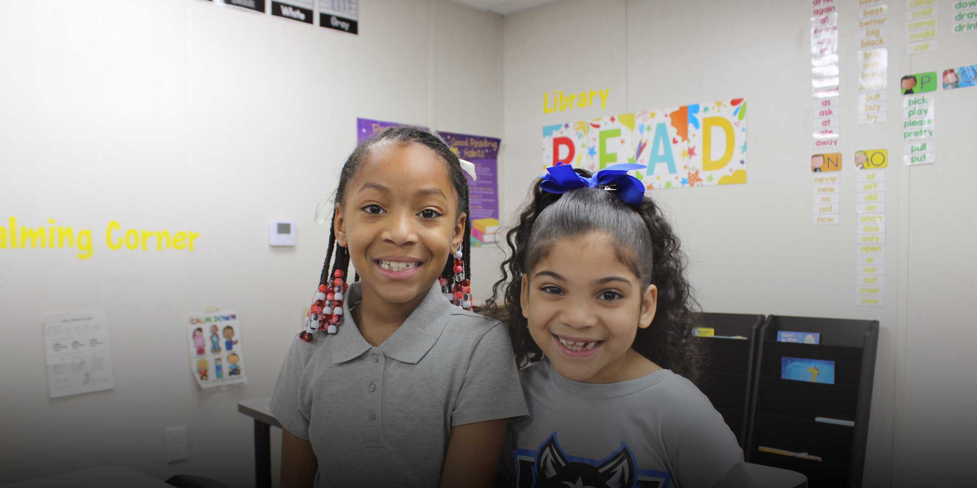 Two smiling students standing together in a classroom reading corner.
